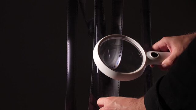 Hand holding a magnifying glass closely inspecting multiple vertical film strips, focusing on details and analyzing the frames against a dark background, representing investigation and research