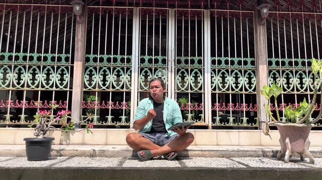 Adult asian man is sitting in front of heritage house in Kotagede while watching online worldcup championship by using an ipad