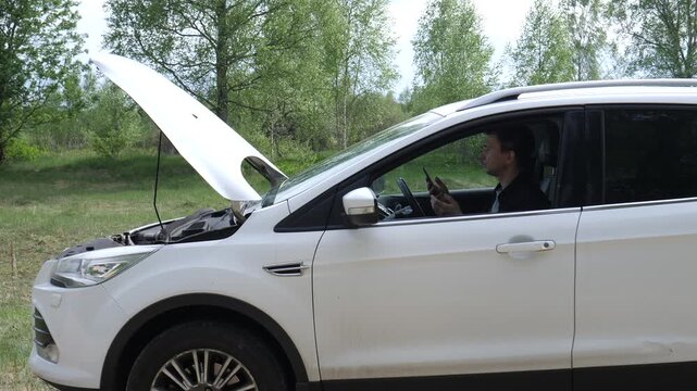 Man sitting inside white suv with hood open on a country road, using mobile phone to call for roadside assistance after vehicle broke down in remote forested area