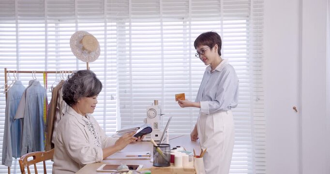 Asian senior female tailor offers POS terminal for cashless checkout while adult customer holds credit card, contactless tap payment inside boutique fashion atelier with sewing tools and workflow