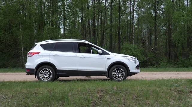 Man standing with the hood open on his broken down white suv by the side of a dirt road, troubleshooting an engine problem with a green forest in the background