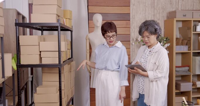 Asian mother and daughter standing together inside boutique atelier small business storage room checking stock boxes with tablet teamwork process family fashion clothing e-commerce preparation