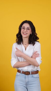 Confident young woman posing with crossed arms and smiling at camera on yellow studio background. Vertical video with copy space, leadership, business, success and modern lifestyle concepts.
