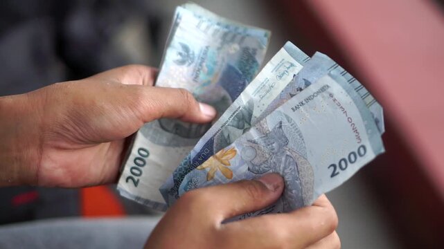 Hands counting Indonesian two thousand rupiah banknotes with a blurred outdoor background.