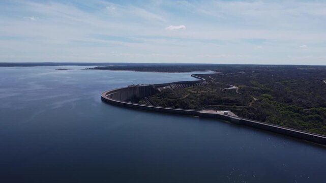 4K high-altitude aerial drone video of the massive Almendra Dam and reservoir in Salamanca, Spain. The wide panoramic shot showcases the impressive curved concrete structure holding back deep blue wat