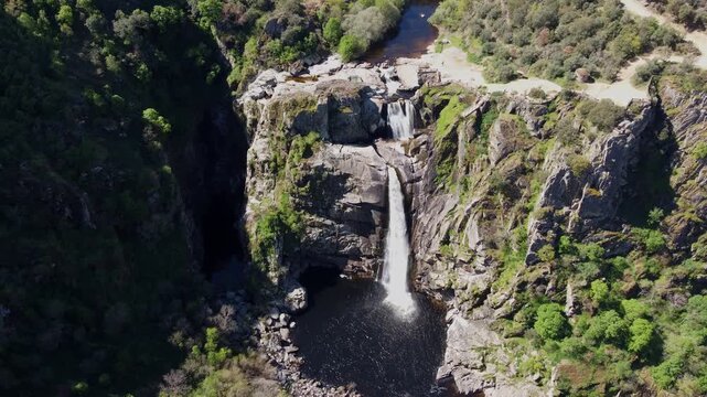 4K aerial drone video pulling back and ascending over the Pozo de los Humos waterfall in Salamanca, Spain. The reveal shot captures the powerful double cascade plunging into a dark pool, surrounded by