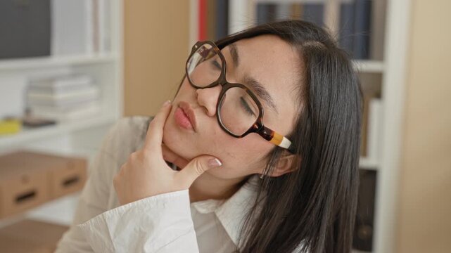Woman wearing tortoiseshell glasses with hand on chin and pensive expression in office with shelves, binders and storage boxes; contemplation.
