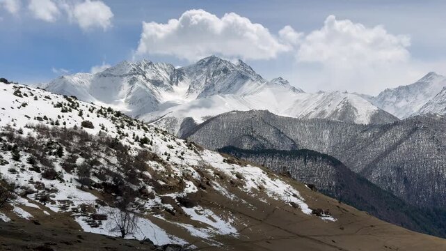 Snow covered mountain peaks rise above mountain slopes beneath clouds across a wide highland landscape. Mountain range.