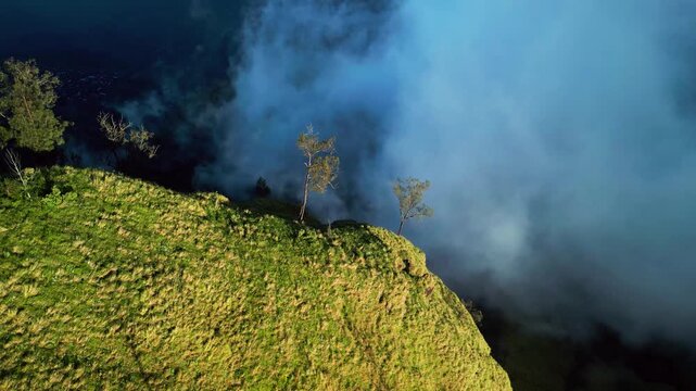 Clouds flowing around steep mountain ridge above volcanic lake, sunlit slopes and grassy cliff edge emerging through drifting fog in aerial tilt-down drone footage