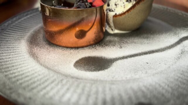 Cinematic close-up of a plated chocolate lava cake in a copper tin, garnished with strawberries and served with vanilla ice cream. The focus is on the artistic powdered sugar stencil of a spoon.