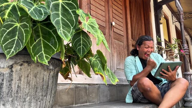 Indonesian sitting down on a Javanese house&rsquo;s terrace, watching football championship online using an ipad during the day