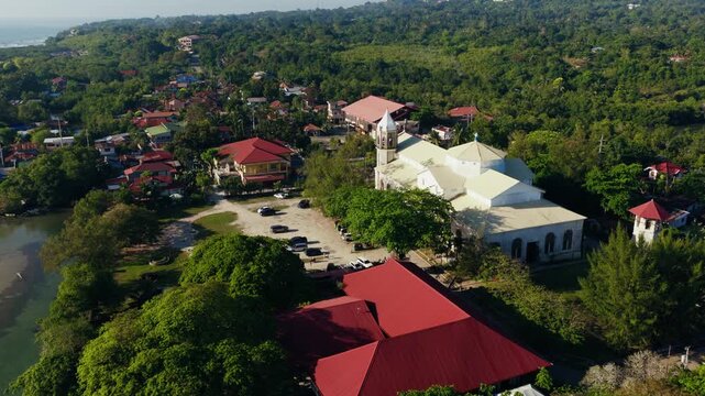 Dauis Church and tropical shoreline beside calm waters in Bohol, drone approach from behind over to mountains