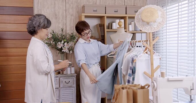Asian senior female mother and adult female daughter teamwork inside boutique atelier small family fashion shop reviewing garment on hanger checking design and quality clothing preparation order