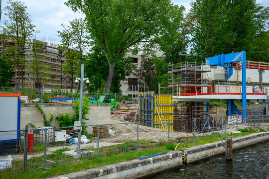 Urban construction site on the banks of the River Spree in Berlin, Germany, showing bridge building work, scaffolding, concrete structures, trees and waterfront development under a cloudy sky.