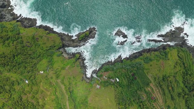 Aerial view of Banyu Tibo Waterfall falling into the ocean with rocky cliffs and crashing waves in Girisubo, Special Region of Yogyakarta, Indonesia.
