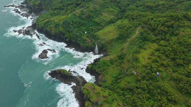 Aerial view of Banyutibo Water Fall cascading into the ocean along the rocky coastline with lush green terraced hills in Girisubo, Special Region of Yogyakarta, Indonesia.