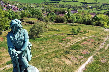 Das monumentale Vercingétorix-Denkmal auf einem steinernen Sockel, eingebettet in eine grüne Hügellandschaft mit Bäumen unter einem strahlend blauen Himmel. © fotoping