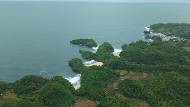 Aerial view of Sedahan Beach featuring lush green hills, terraced fields, and a rocky coastline with sandy coves in Girisubo, Special Region of Yog, Indonesia.