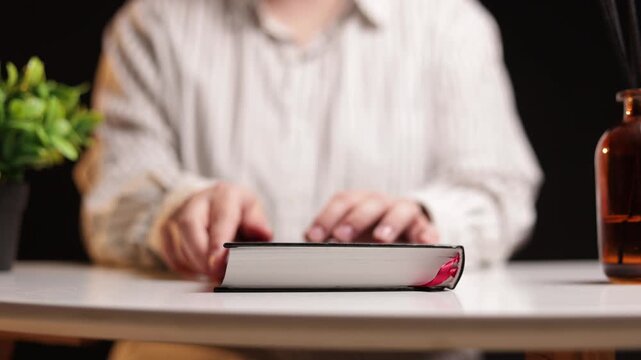 Person closing a book with a red ribbon bookmark on a table