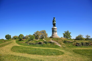 Das monumentale Vercingétorix-Denkmal auf einem steinernen Sockel, eingebettet in eine grüne Hügellandschaft mit Bäumen unter einem strahlend blauen Himmel. © fotoping