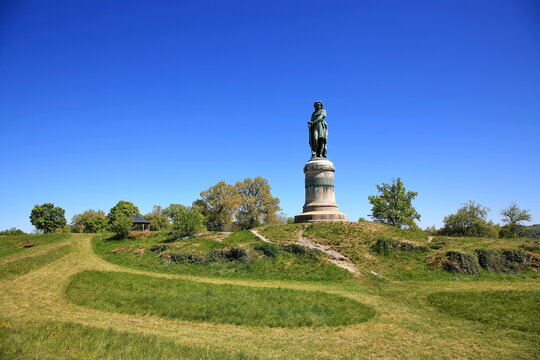 Das monumentale Vercing&eacute;torix-Denkmal auf einem steinernen Sockel, eingebettet in eine gr&uuml;ne H&uuml;gellandschaft mit B&auml;umen unter einem strahlend blauen Himmel.