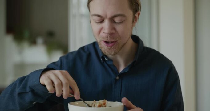 Caucasian man eats oatmeal with fruits. Male eating breakfast at kitchen at home. 4K 10-bit