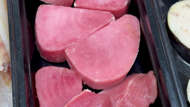 Pink tuna steaks on black tray, supermarket shelf closeup, chilled display of raw fresh fillets arranged in neat rows, subtle