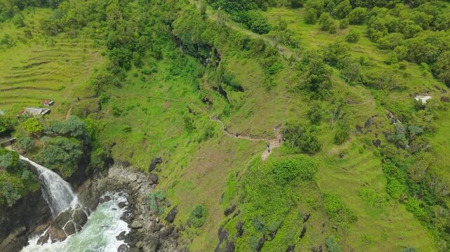 Aerial view of Banyutibo Waterfall cascading into the sea from lush green cliffs with terraced fields and a walking path in Girisubo, Special Region of Yogyakarta, Indonesia.