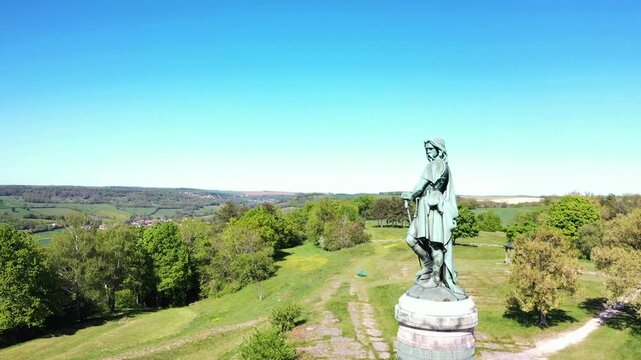 Das monumentale Vercing&eacute;torix-Denkmal auf einem steinernen Sockel, eingebettet in eine gr&uuml;ne H&uuml;gellandschaft mit B&auml;umen unter einem strahlend blauen Himmel.