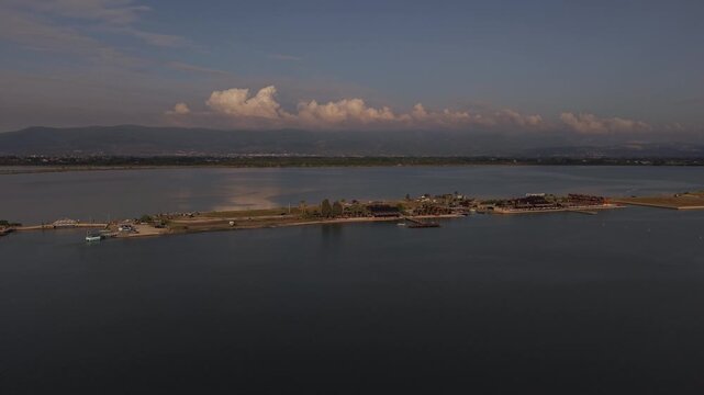 Wide angle drone footage of Patok Lake, Albania, showing calm lagoon waters, a narrow shoreline, distant mountains, dramatic clouds, and peaceful coastal scenery.
