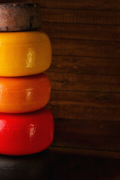 Several types of cheese are stacked on top of each other on a dark background. Cheese wheels in latex. Semi-hard and hard cheeses. Mature cheeses in packaging. Tilsiter, Gouda, Parmesan, Cheddar, Mimo