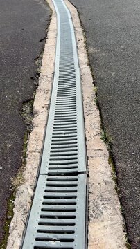 Galvanized drainage grate along stadium curb, long metal channel between asphalt and concrete, repeating slot pattern, leading