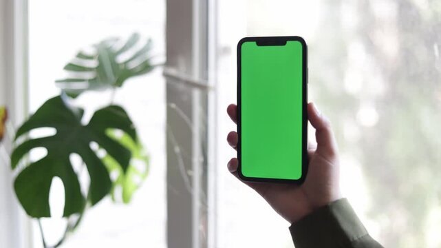 Person holding a smartphone with a green screen in front of a window with plants