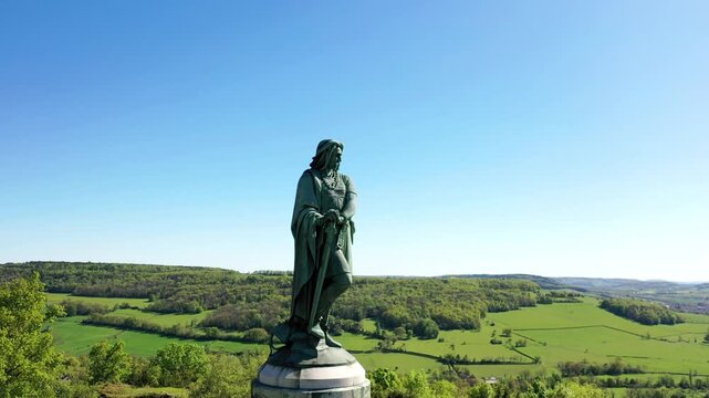 Das monumentale Vercing&eacute;torix-Denkmal auf einem steinernen Sockel, eingebettet in eine gr&uuml;ne H&uuml;gellandschaft mit B&auml;umen unter einem strahlend blauen Himmel.