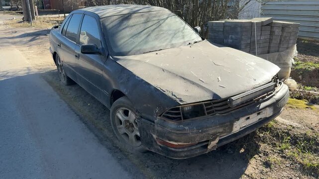 Dusty black sedan abandoned by roadside, frontend collision damage with cracked bumper, tinted windows covered in grime