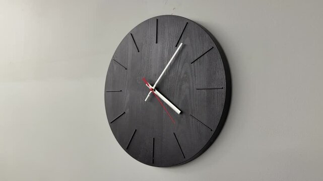 Black wooden wall clock on grey wall, minimalist timepiece with textured round face, white hour and minute hands, red second hand