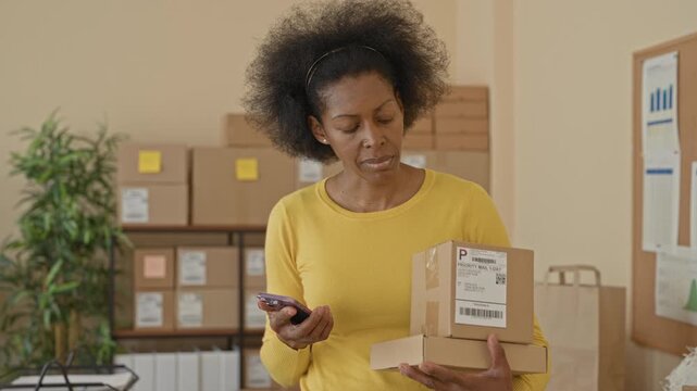 Woman holding parcels and a smartphone, checking shipping labels and her phone while balancing boxes and standing by shelves in a building; focused small business.