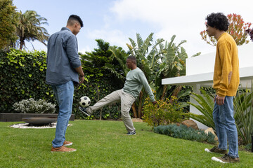 Diverse male friends playing soccer on backyard lawn, kicking ball near fire pit wearing sneakers
