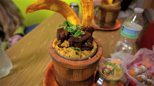 A close-up shot of a delicious mofongo dish made of mashed green plantains, garlic, and chicharron, served in a traditional wooden mortar and pestle at a restaurant.