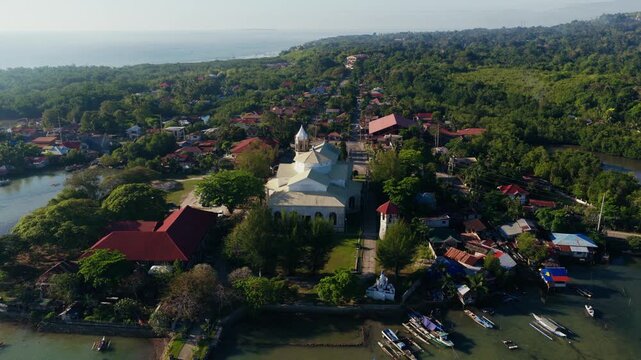 Dauis Church surrounded by tropical homes and coastal shoreline, drone rearview orbit
