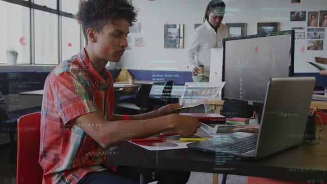 Seated man scanning printout at table, typing on laptop for business while colleagues passing