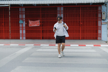 Fototapeta premium Young man walking on a pedestrian crosswalk in an Asian city, using a smartphone, embodying urban connection, street style, technology, and modern outdoor leisure