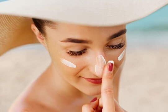 Young woman smiling while applying protective white sunscreen cream to her face at a beautiful beach during a bright summer day