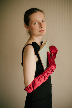 Elegant woman with frecklesin black dress and red gloves reading book in vintage interior with warm light.