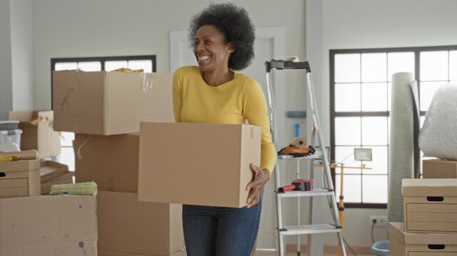 Woman carries cardboard box while smiling and lifting inside building filled with stacked boxes and packing materials; moving day joy.
