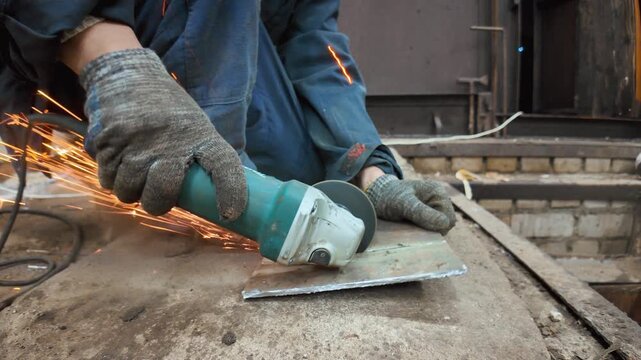 Professional craftsman in protective gloves using an angle grinder for cutting a metal sheet, with bright sparks flying during the industrial metalworking process in a workshop
