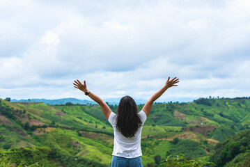 Fototapeta premium Back view of a freedom woman raising arms to the sky on top of a mountain. Happy traveler enjoying beautiful green nature landscape of rolling hills and mountains under a cloudy sky.