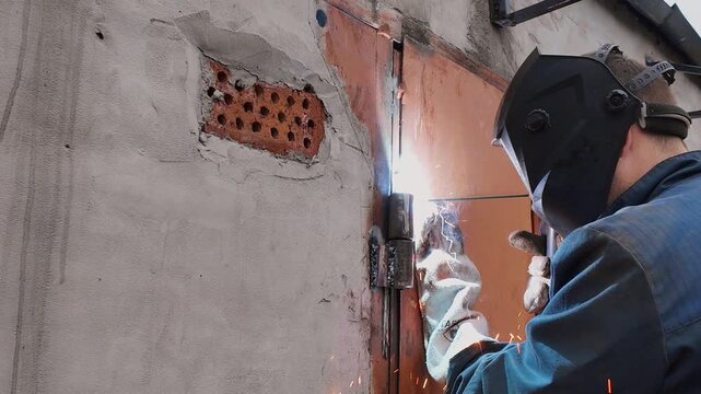 Skilled worker in protective gear performing electric arc welding on a garage door hinge, with bright sparks flying as he precisely joins the metal parts on a concrete and brick wall