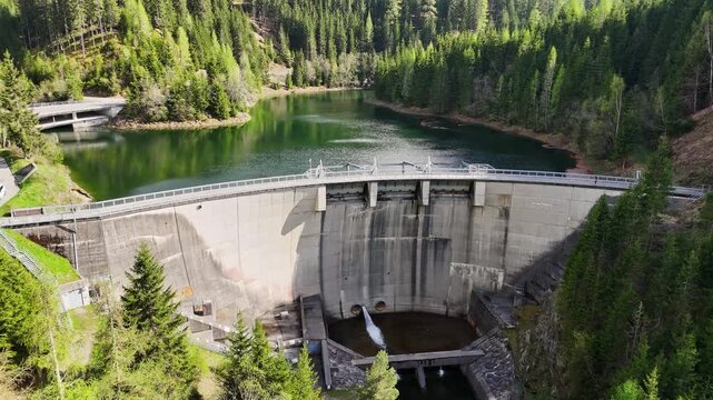 Aerial view of Kraftwerk Bodendorf and Sperre Paal dam wall with a green reservoir surrounded by dense evergreen forests in Murau, Styria, Austria.