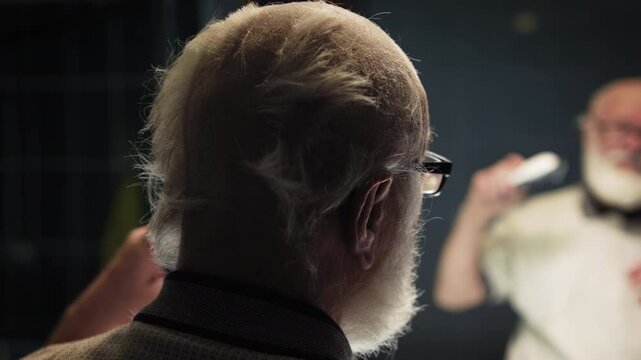 Elderly man in glasses monitors his haircut with a trimmer in front of a mirror